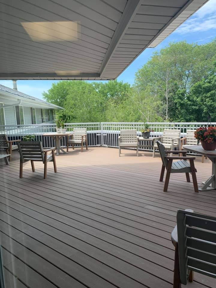 A spacious outdoor deck with tables and chairs, sheltered by an awning, surrounded by lush greenery.