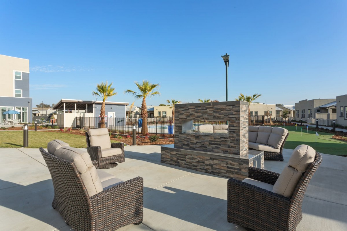 A sunny outdoor patio area with a modern fireplace and comfortable seating surrounded by palm trees.