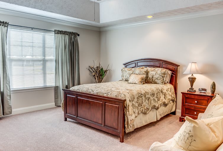 A cozy bedroom featuring a wooden bed frame, floral bedding, and natural light from a large window with curtains.