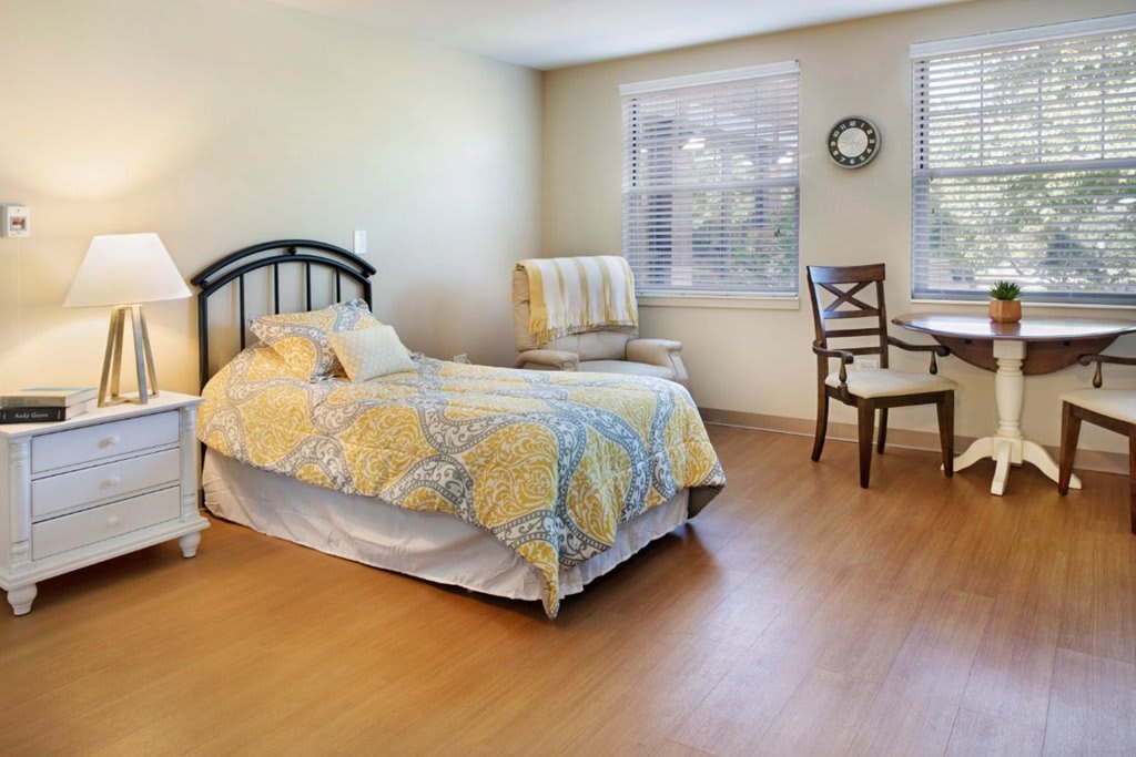A cozy bedroom featuring a single bed with a patterned quilt, a chair and table set by the window, and bright natural lighting.