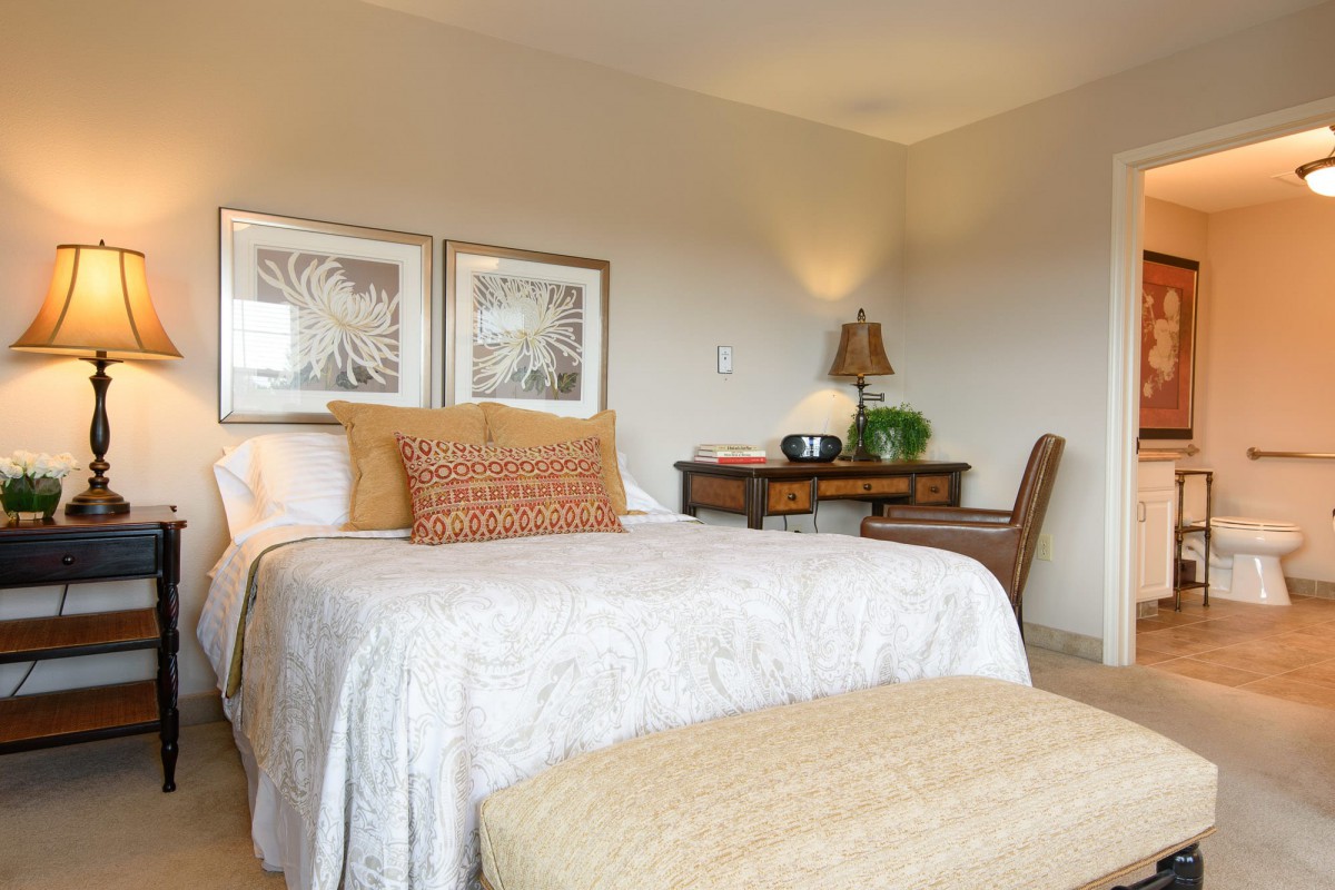 A cozy bedroom with a neatly made bed, decorative pillows, and a desk with a lamp.