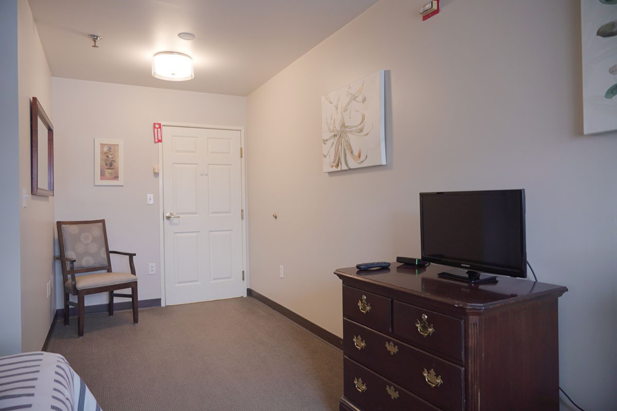A simple room featuring a dresser with a television, a chair, and a door with wall art.