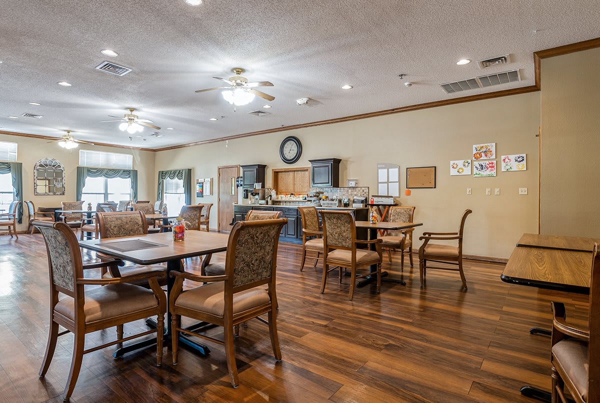 A spacious dining area with wooden tables and chairs, featuring ceiling fans and a nearby kitchen space.