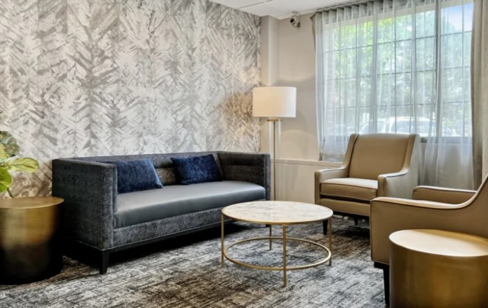 A modern living room featuring a dark gray sofa, an armchair, a round coffee table, and a tall floor lamp, set against a textured wall and large window with curtains.