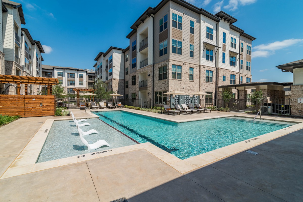 A modern residential apartment complex featuring an outdoor swimming pool under a clear blue sky.