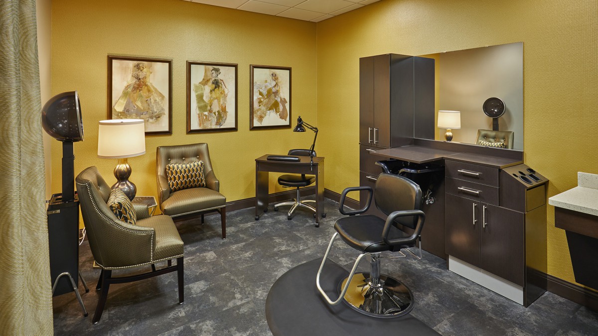 A modern salon interior featuring a styling chair, desk, artwork on the walls, and a small waiting area with comfortable seating.