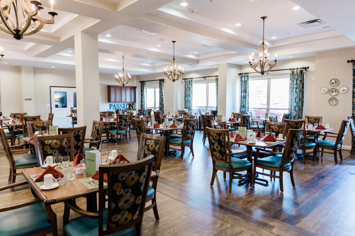A spacious and elegantly decorated dining area with several tables and chairs, featuring chandeliers and natural lighting.