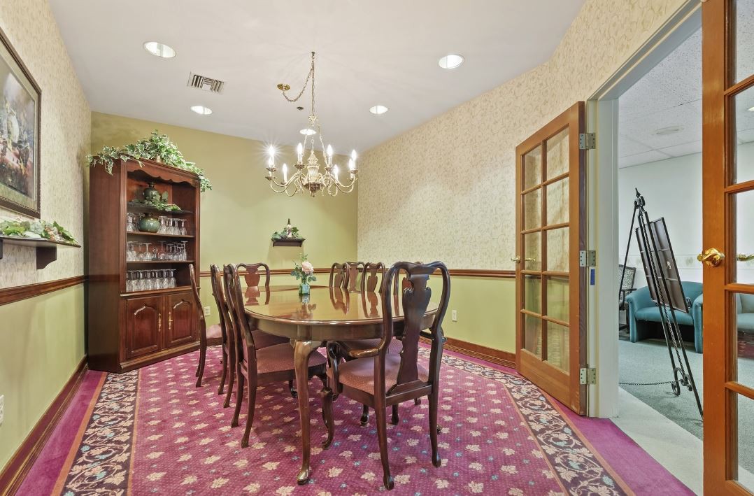 A formal dining room with an elegant wooden table and chairs, a chandelier overhead, and a patterned carpet underfoot.