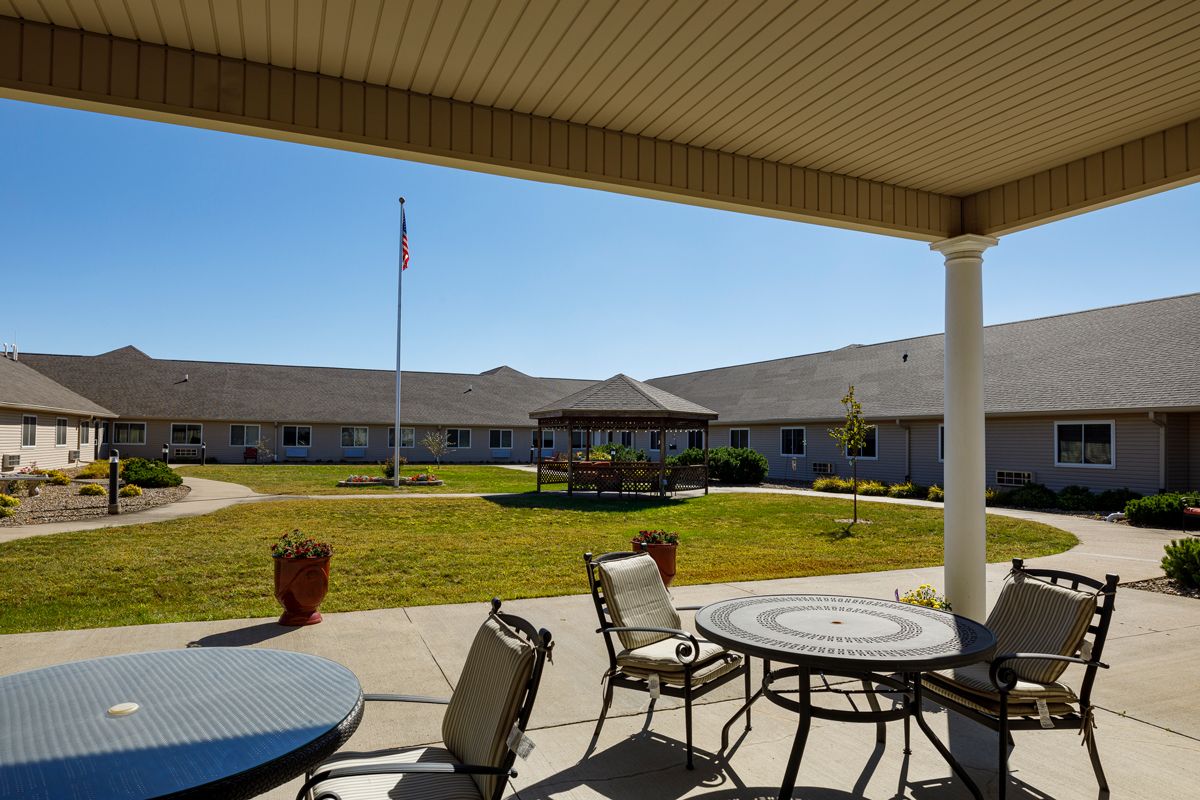 A view from a covered patio with outdoor seating, overlooking a building and a well-maintained lawn under a clear blue sky.