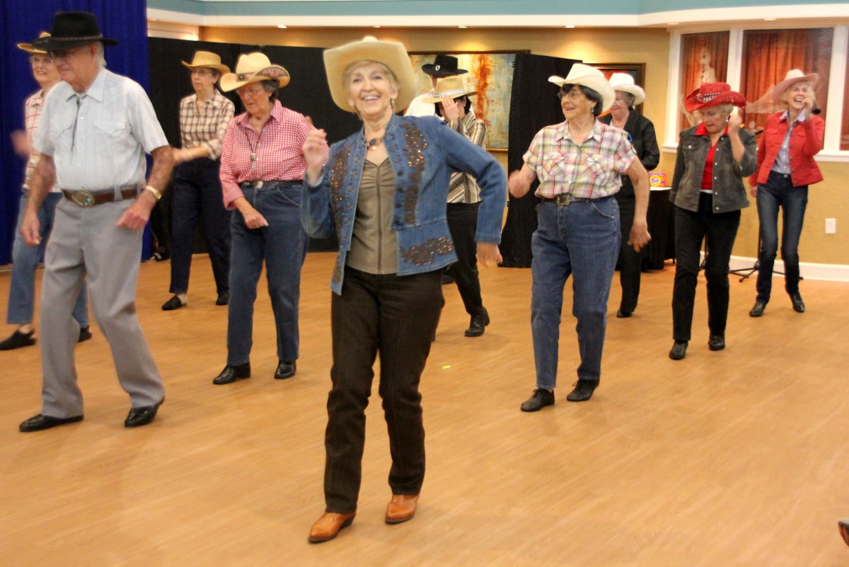 A group of people line dancing indoors, dressed in country-style outfits with cowboy hats.
