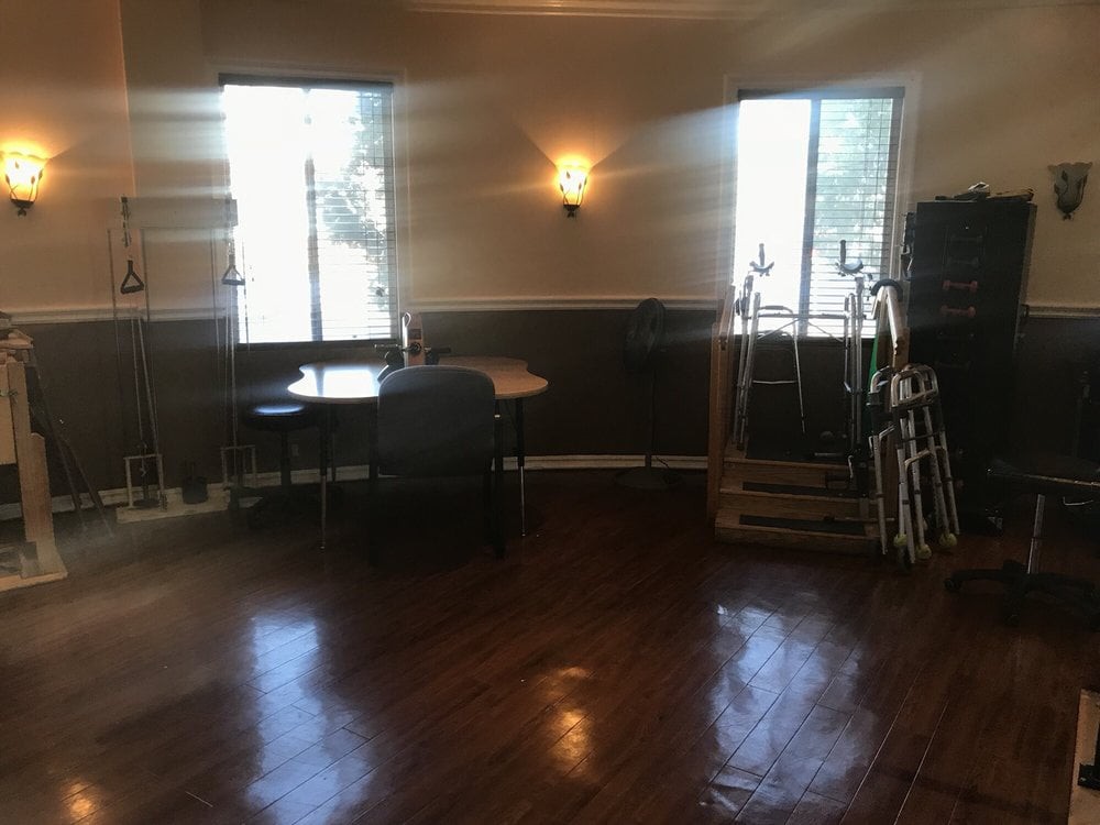 An empty room with wooden floors, a few tables and chairs, and two windows letting in natural light.