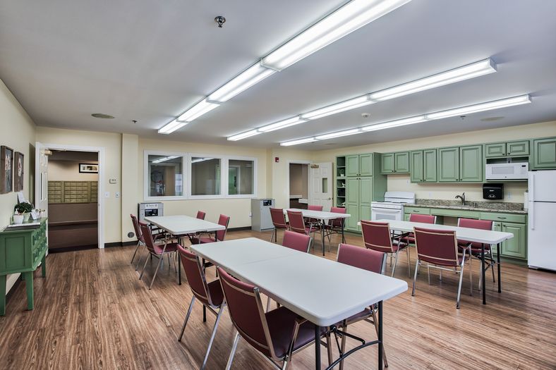 A well-lit classroom with tables and chairs, featuring a kitchen area in the background.