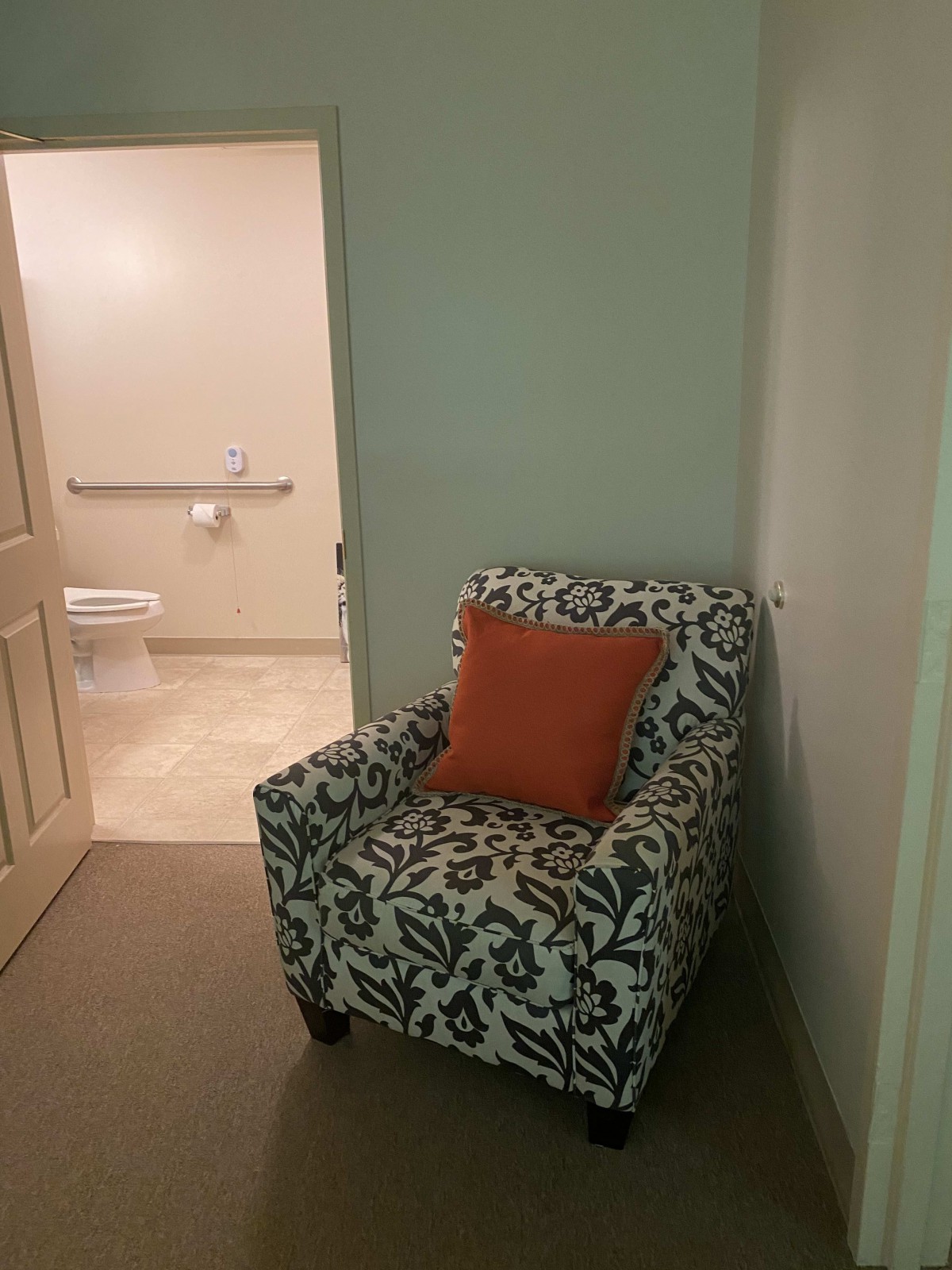 An armchair with a decorative pillow sits near a hallway leading to a bathroom.