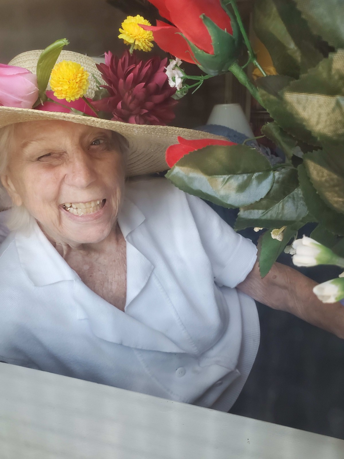 An elderly woman wearing a hat, surrounded by flowers, with a joyful smile.