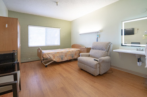 A comfortable and minimalistic hospital room featuring a patient bed, a recliner chair, and large windows that allow natural light.