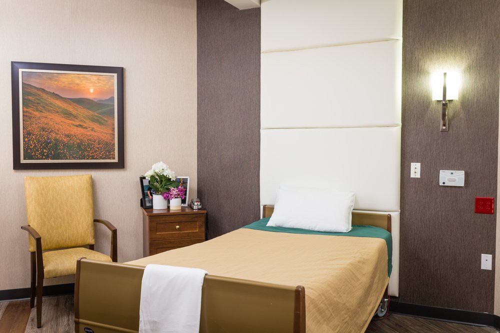 A tidy hospital room with a single bed, chair, and nightstand adorned with flowers.