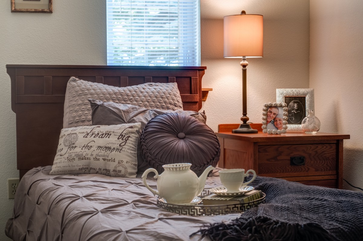 A cozy bedroom scene featuring a neatly made bed with decorative pillows, a tea set on a tray, and a softly lit lamp on the nightstand.