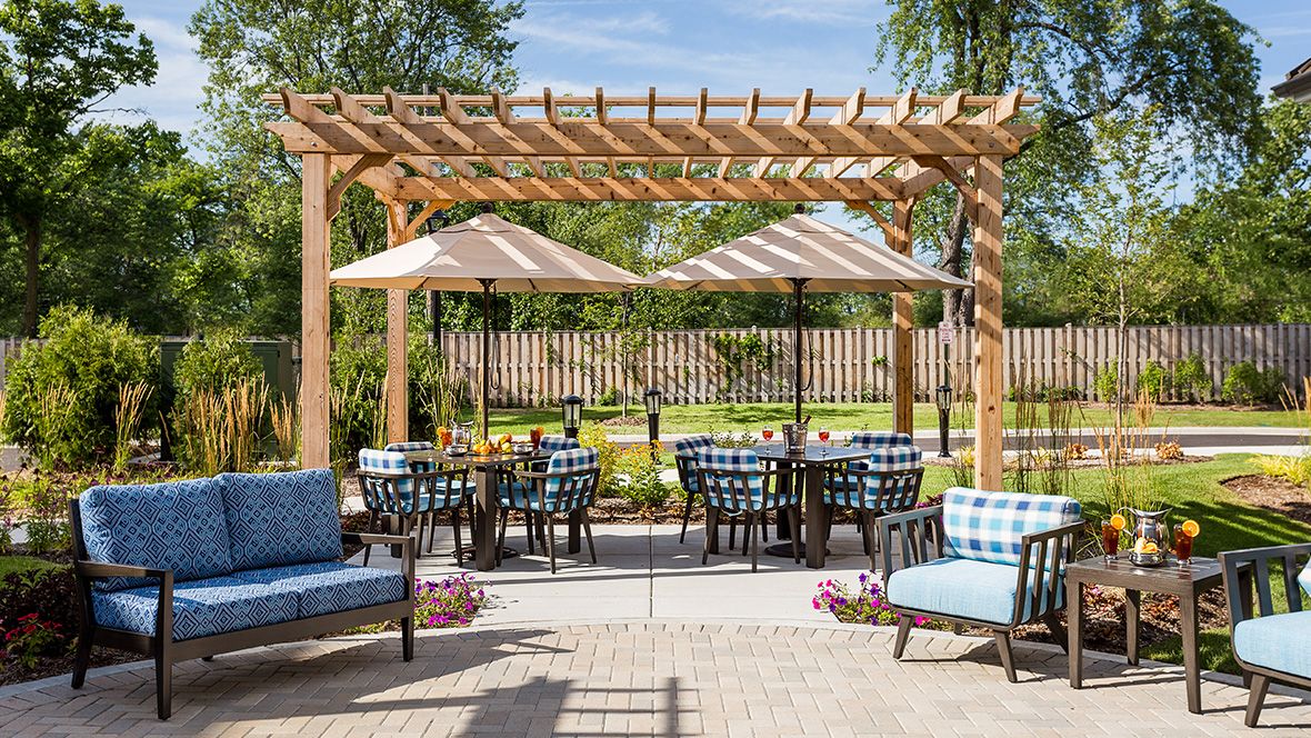 A cozy outdoor patio with comfortable seating under a wooden pergola, featuring umbrellas and surrounded by lush greenery.