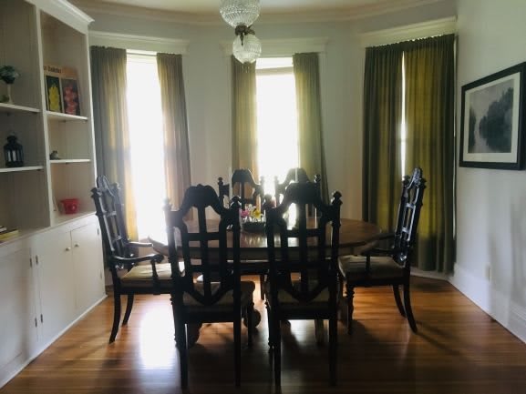 A cozy dining room with a wooden table surrounded by chairs, light filtering through curtains covering large windows.