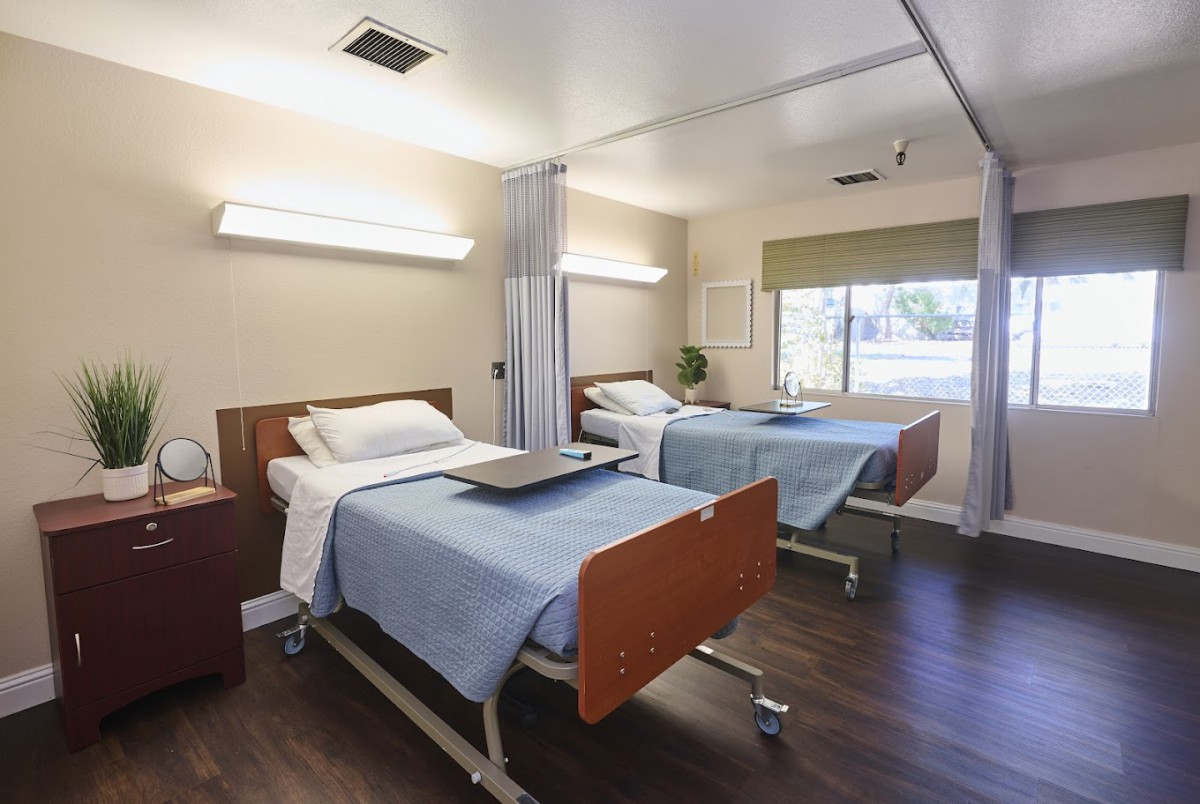 A hospital room with two neatly made beds, each equipped with a small table and situated in a brightly lit space.