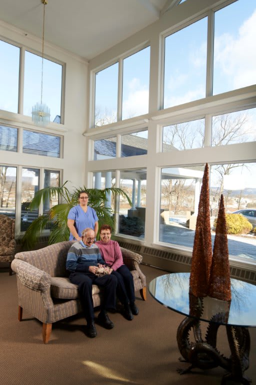 A nurse posing with two senior individuals in a care facility's lounge area.