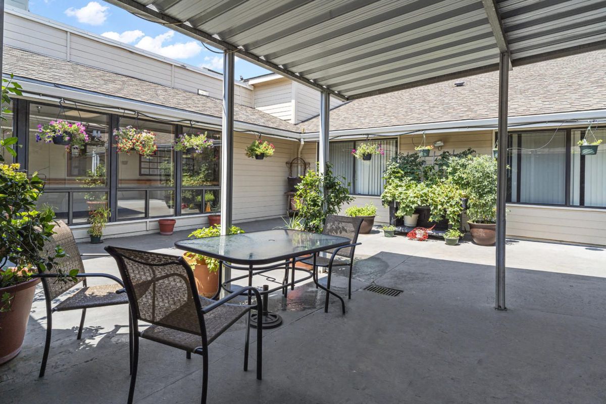 A covered patio area with a glass table and chairs, surrounded by potted plants and hanging baskets under sunny skies.