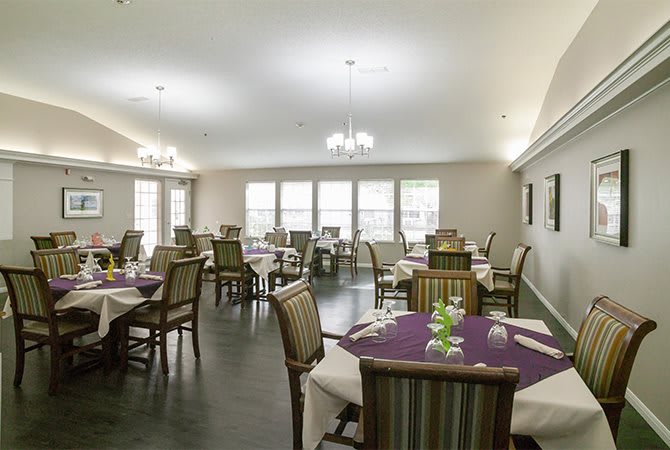 A well-lit dining room with neatly arranged tables and chairs, featuring chandeliers and large windows.