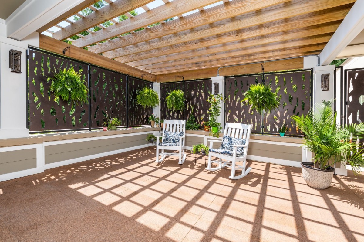 A serene patio area with a pergola providing shade over two white rocking chairs, surrounded by lush hanging plants.