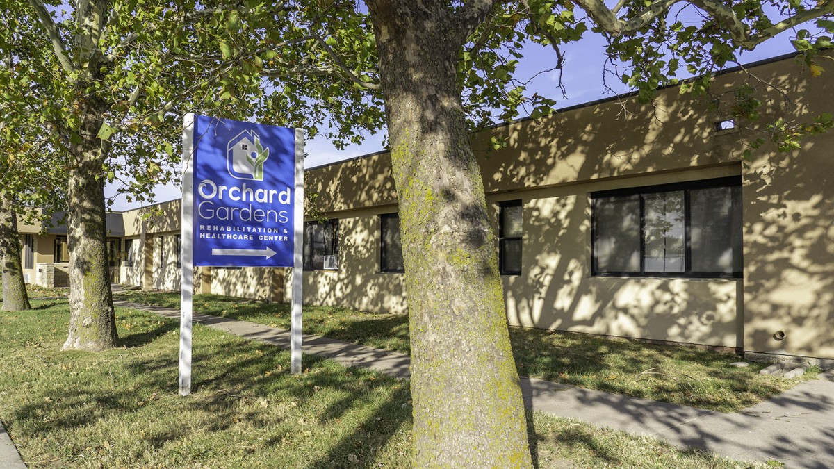 A blue sign stands next to a tree in front of a beige building.