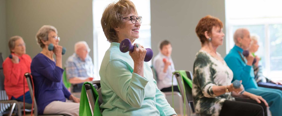 An elderly woman is exercising indoors, lifting a purple dumbbell and smiling.