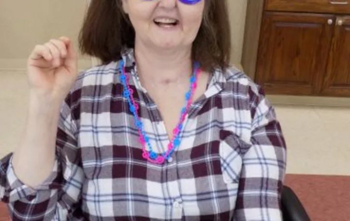 A person wearing a plaid shirt and colorful necklaces is smiling indoors.