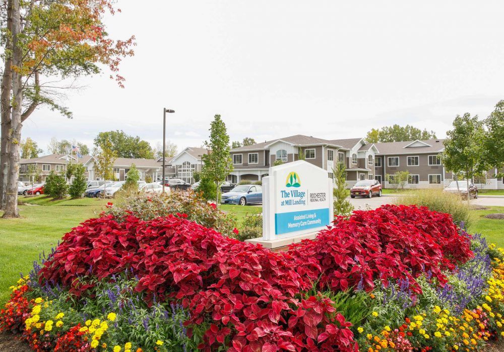 A landscaped area with red flowers in front of a building and a parking lot, featuring a sign.