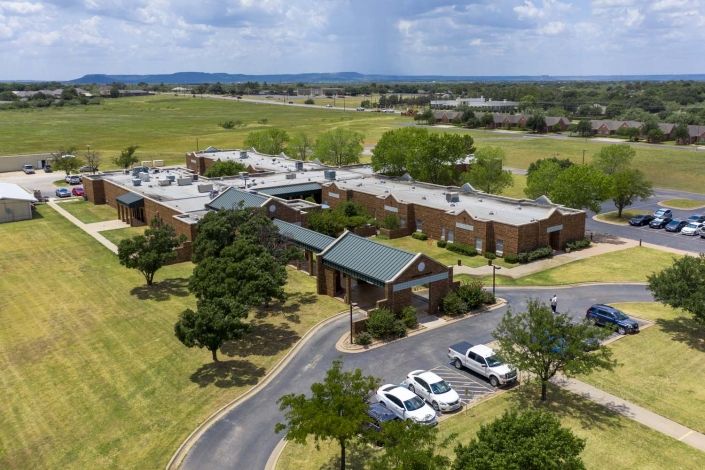 Aerial view of a hospital surrounded by greenery and fields, with a parking lot in front.