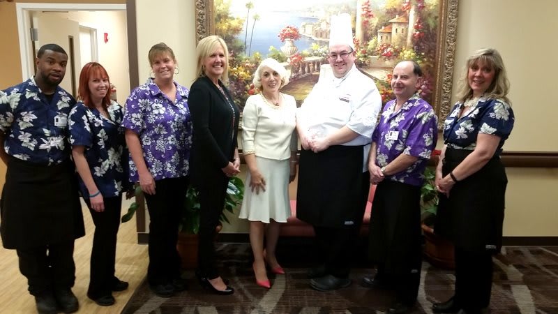 A group of people, including a chef, pose indoors in front of a scenic painting.