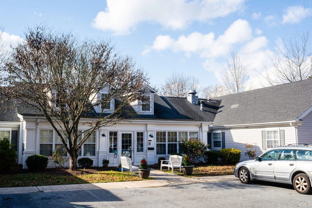 A suburban house with a driveway and parked car, surrounded by trees under a partly cloudy sky.