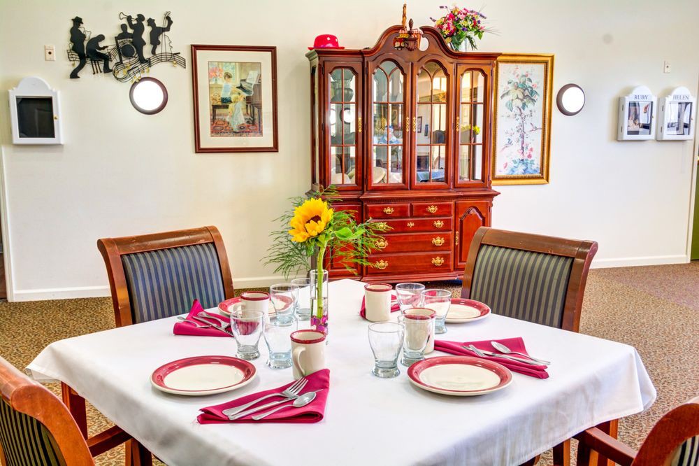 A neatly arranged dining table with red and white dinnerware, surrounded by chairs, a flower vase centerpiece, and a china cabinet in the background.