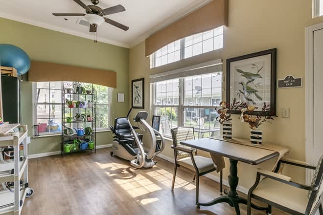 A bright home office with exercise equipment, featuring large windows, a treadmill, a table and chairs, indoor plants, and wall artwork.