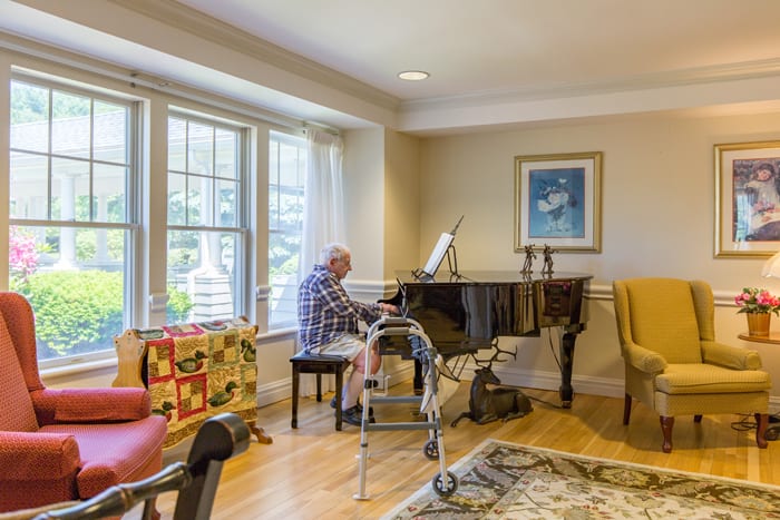 An elderly person playing the piano in a cozy living room with a walker nearby.