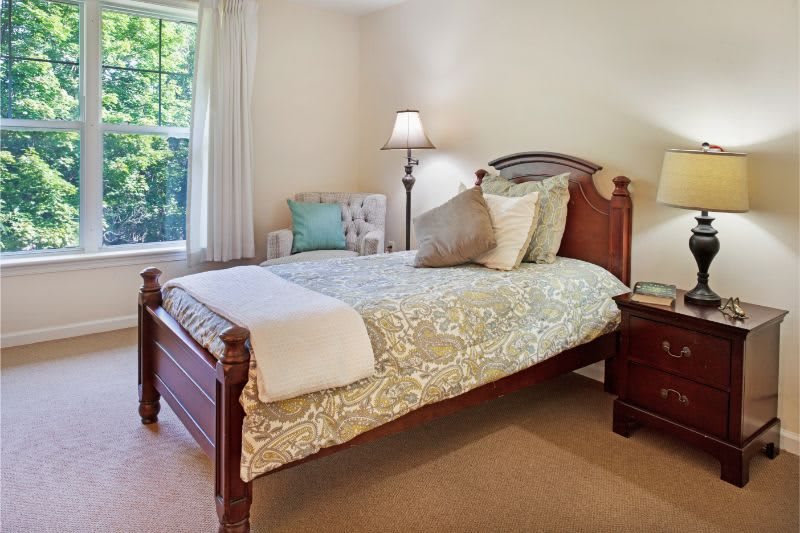 A cozy bedroom with a wooden bed frame and decorative pillows, illuminated by a nearby lamp next to a window.
