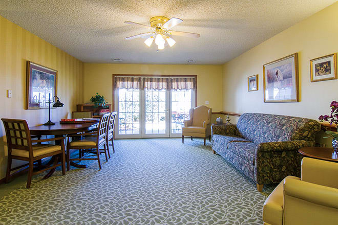 A cozy living room with a sofa, chairs, and a dining table, featuring large windows with curtains.