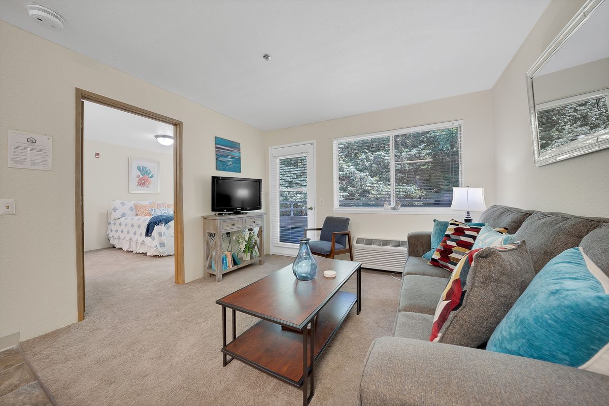 A cozy living room featuring a sofa, coffee table, and TV, with large windows providing natural light.