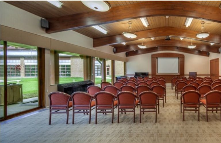 An empty conference room with rows of chairs facing a projection screen, featuring large windows and a carpeted floor.