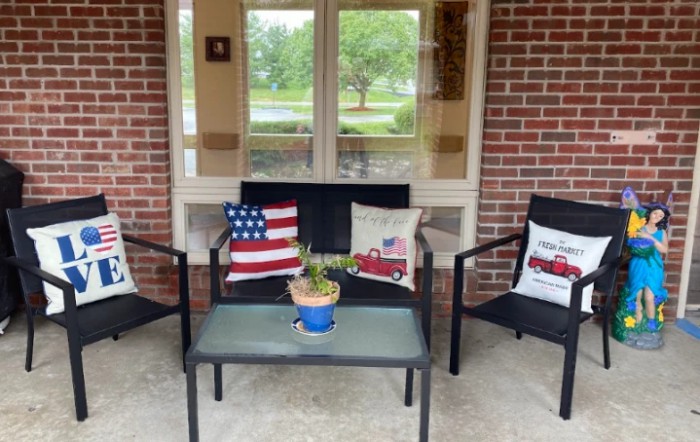 A cozy patio setup with chairs and a table, decorated with American flag and fire truck themed cushions, alongside a potted plant.