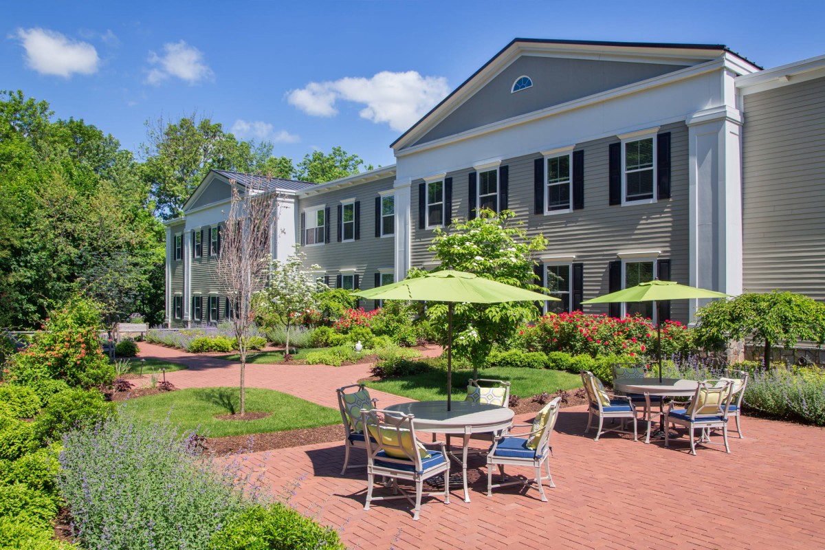 A well-maintained outdoor patio area with tables and chairs, surrounded by lush greenery in front of a large, colonial-style building.