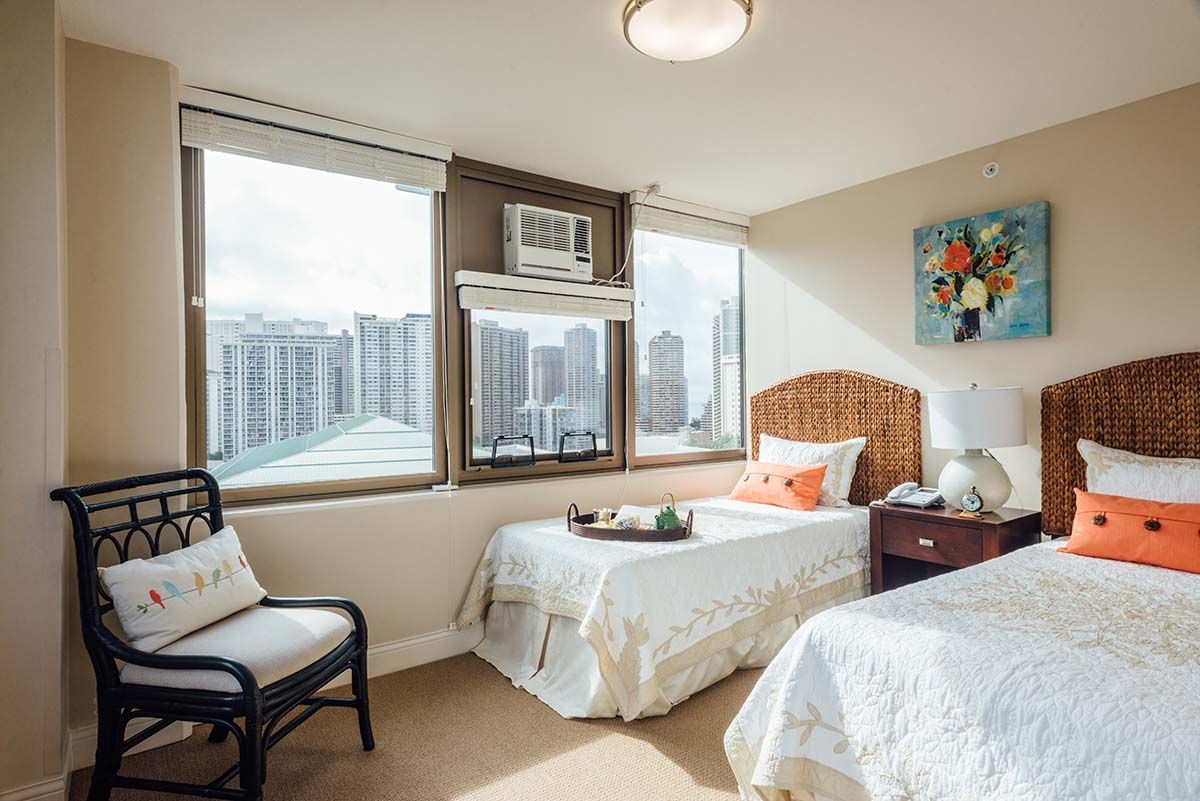 A bright bedroom with twin beds and a view of the city skyline through large windows.