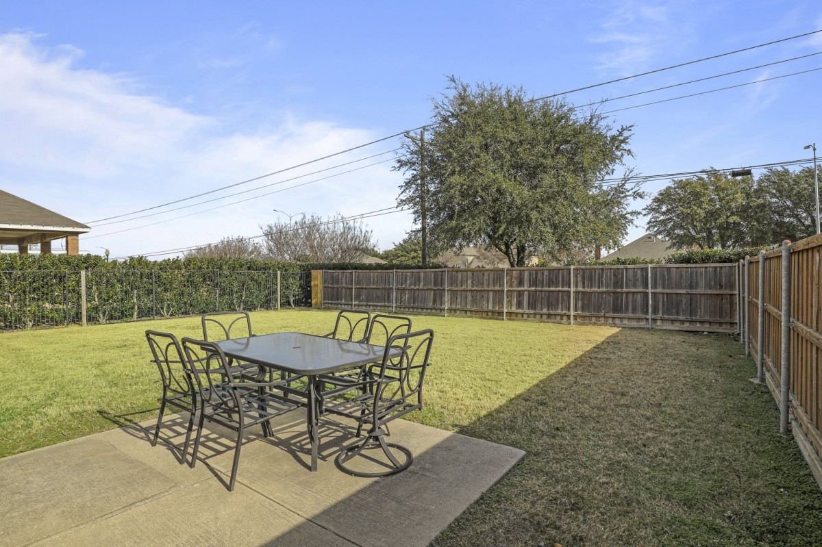 A backyard patio with a table and chairs overlooking a lawn surrounded by a fence.