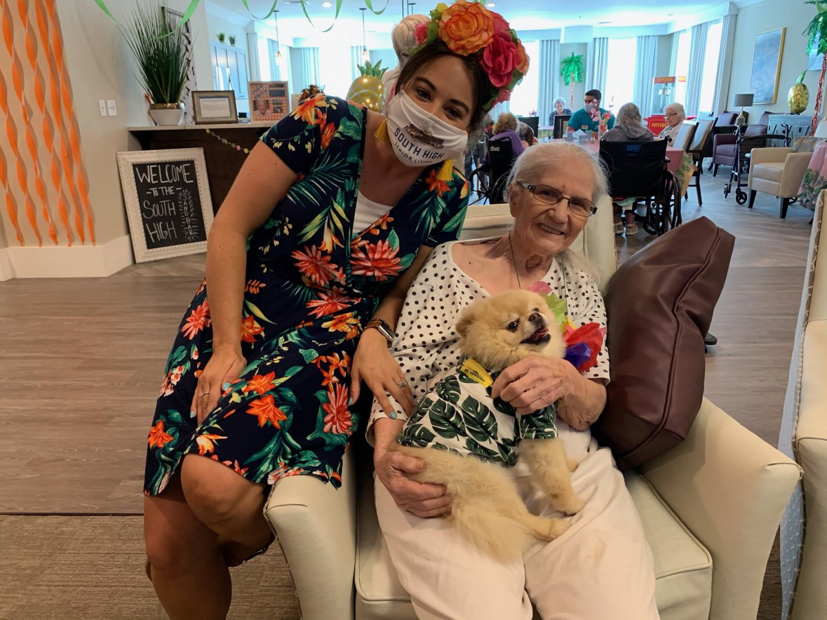 An elderly woman sitting with a small dog on her lap, accompanied by a nurse wearing a floral dress and mask.