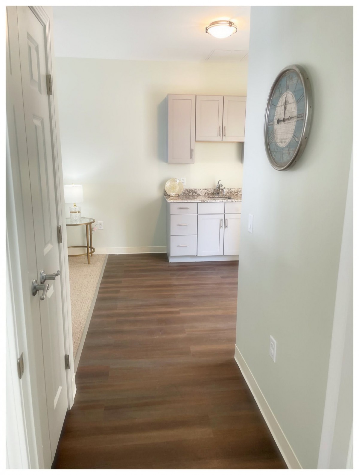 A view of a modern hallway leading to a kitchenette area with wooden flooring and a wall clock.