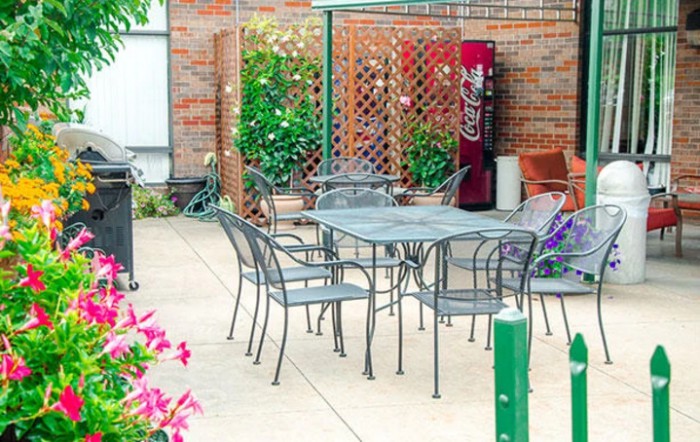An outdoor patio area with metal table and chairs, surrounded by plants and a vending machine in the background.