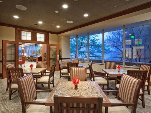 A cozy dining area with neatly arranged tables and chairs, illuminated by ceiling lights and large windows.
